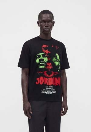 Osoba ubrana w czarny T-shirt z czerwonymi i zielonymi graficznymi twarzami oraz dużym czerwonym napisem "JORDAN", stojąca na jednolitym białym tle.