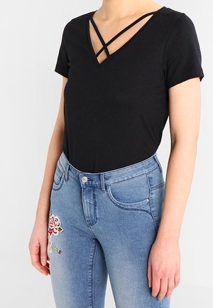 Frau trägt ein schwarzes V-Ausschnitt-Kurzarmshirt mit überkreuzten Trägern und hellblaue Jeans mit Blumenstickerei am Oberschenkel.