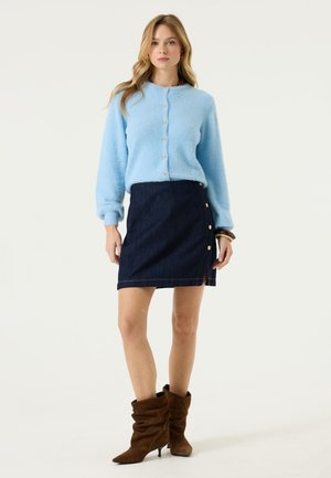 Femme debout, portant un cardigan bleu clair à boutons, une jupe en jean foncé avec des boutons sur le côté et des bottes marron souples à talon, sur fond blanc.