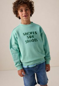 Sweat-shirt turquoise avec "SHORTS SUN SHADES" en texte vert, associé à un short en denim. Tissu doux, coupe décontractée, poignets et ourlet côtelés.