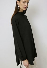 Zwarte longsleeve top met een hoge kraag, voorzien van zijsplitten en verstelbare strikjes aan de zoom. Glad textuur en relaxed fit.
