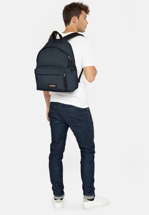 Zaino blu scuro con tessuto testurizzato, spallacci regolabili, tasca frontale e zip, indossato da una persona con una camicia bianca e jeans.