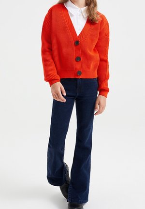 Osoba ubrana w jasnopomarańczowy dzianinowy kardigan z dużymi guzikami, białą koszulę z kołnierzykiem, ciemnoniebieskie dzwony i czarne buty na białym tle.