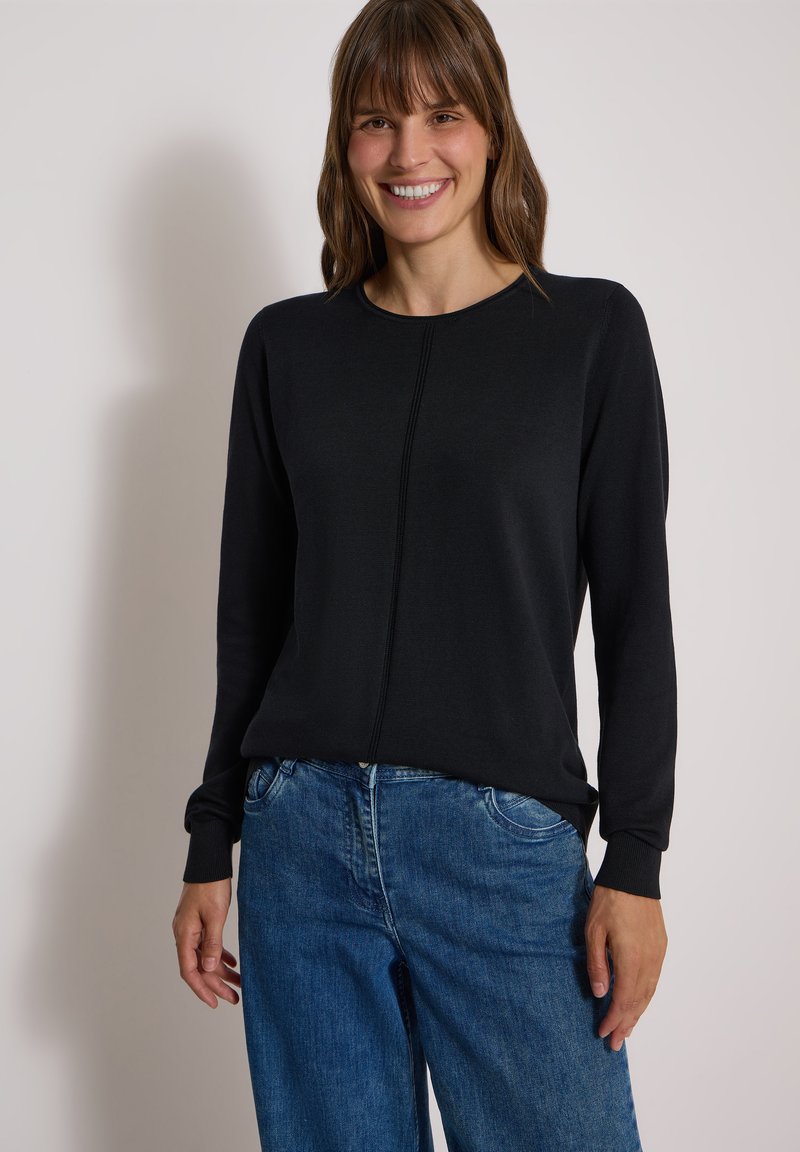 Femme souriante aux cheveux bruns, portant un shirt noir à manches longues et un jean bleu, debout devant un fond clair uni.