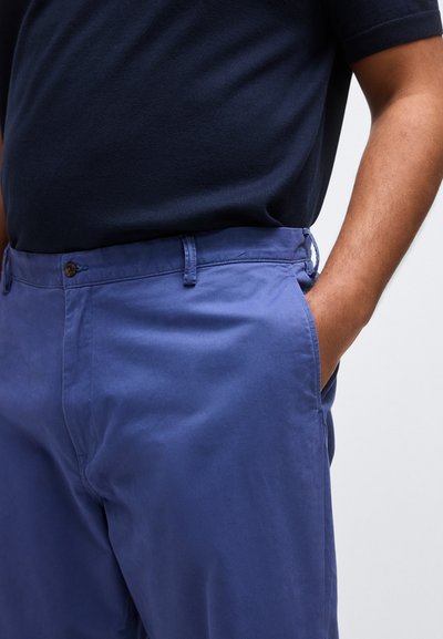 Άνδρας που φοράει μπλε ναυτικό πουκάμισο και ινδικό μπλε παντελόνι με το ένα χέρι χαλαρά τοποθετημένο στην τσέπη, σε απλό ανοιχτό φόντο.