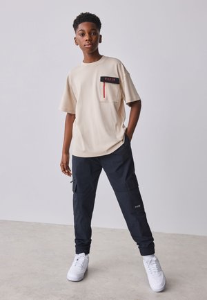 Tienerjongen staat met één hand in zijn zak, draagt een beige T-shirt met borstzak en zwarte cargobroek met witte sneakers.