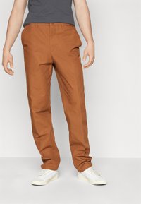 Brown cotton trousers with a straight leg design, featuring side pockets and subtle branding. Paired with white sneakers.