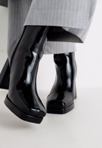 Botine negre din piele lăcuită, cu toc gros, vârf pătrat și textură netedă. Asortate cu pantaloni gri cu dungi.