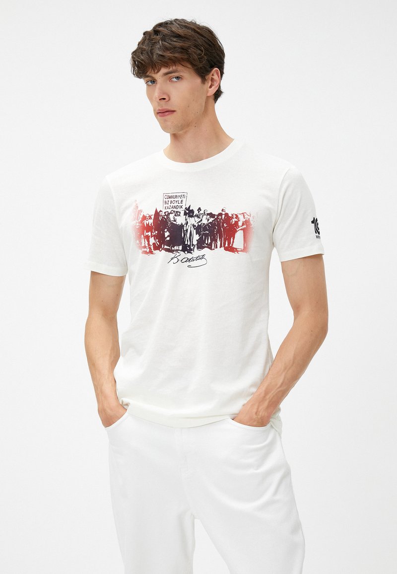 Camiseta blanca de algodón con un estampado gráfico de figuras históricas en negro y rojo, con una firma debajo. Mangas cortas y cuello redondo.