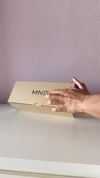Brun papkasse med teksten "MNG" på siden, holdt af en hånd, placeret på en hvid overflade mod en lyserød væg.