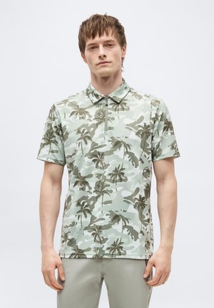 Mladić u kratkoj svijetlozelenoj polo majici s uzorkom palmi i svijetlim hlačama, stoji ispred jednobojne pozadine.