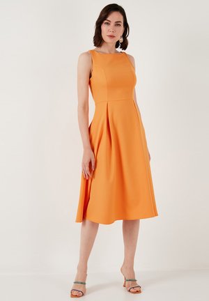 Robe sans manches orange avec un corsage ajusté, une jupe évasée et des plis. Texture lisse, design mi-long, accompagnée de sandales à lanières.