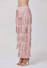 Jupe maxi frangée rose en tissu doux, avec des sections superposées et une taille élastique. Portée avec des chaussures à talons blancs.