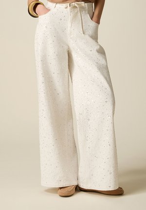 Pantalon blanc à jambes larges avec motif en sequins argentés, taille à cordon, mains dans les poches, associé à des chaussures plates marron et des bracelets dorés.