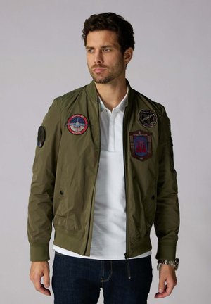 Blouson bomber vert olive avec des poignets côtelés, fermeture éclair à l'avant et plusieurs écussons brodés sur la poitrine et les manches ; en nylon léger.