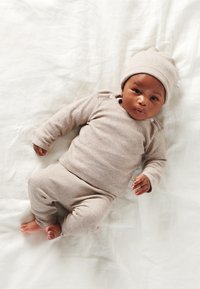 Hellbrauner, gerippter Strickanzug für Babys mit langen Ärmeln, passenden Hosen und einer Mütze, auf einem weißen Stoffhintergrund ausgelegt.