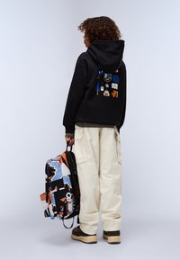 Felpa nera con toppe grafiche colorate sulla schiena, abbinata a pantaloni cargo color crema e uno zaino multicolore con un motivo.