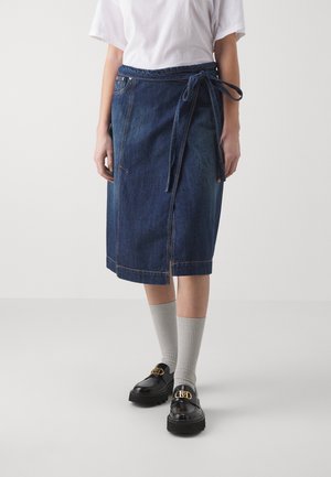 Persona che indossa una gonna avvolgente in denim scuro, lunga fino al ginocchio, con cintura legata, camicia bianca, calze grigie a costine e moccasini neri con dettagli in fibbia dorata.