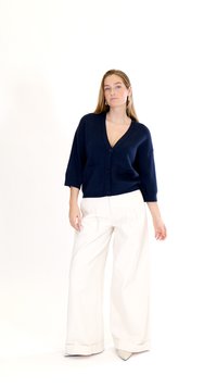 Žena stoji, nosi tamnoplavi kardigan s gumbima i džepovima te široke bijele hlače, na jednobojnoj bijeloj pozadini.