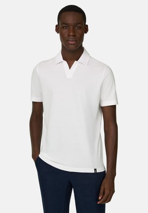 Jeune homme portant un polo blanc à manches courtes et un pantalon bleu foncé, debout avec une main dans la poche sur un fond uni.