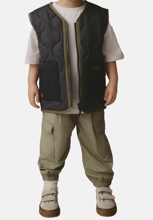 Veste matelassée noire avec bordure olive, fermeture éclair et poches, associée à un pantalon cargo vert et des baskets beige avec des accents de lanières.