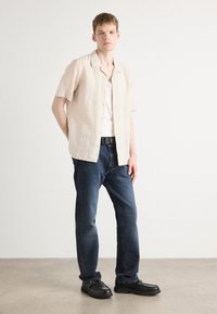 Light beige short-sleeve shirt, button-up design, worn over a white tank top. Dark blue jeans and black shoes with a textured sole.