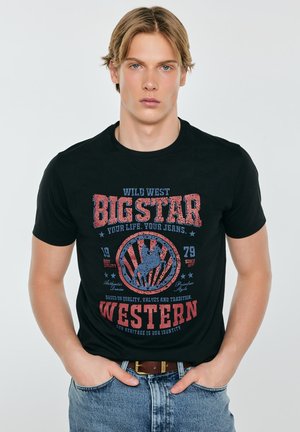 Młody mężczyzna o blond włosach, ubrany w czarny T-shirt z czerwoną i niebieską grafiką w westernowym stylu oraz jasnoniebieskie dżinsy, ręce w kieszeniach.