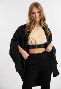 Sort oversized skjorte med lange ærmer lagdelt over en beige cropped top. Højtaljede sorte bukser med sidelukkere. Glatte teksturer.