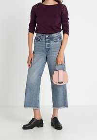 Kobieta w fioletowej bluzce z koronkowymi rękawami, w niebieskich dżinsach z wysokim stanem, czarnych butach, trzymająca małą różową torebkę za uchwyt.