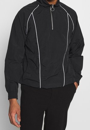 Personne portant une veste-pull noire avec des détails en passepoil blanc et un col zippé jusqu'à mi-hauteur, associée à un pantalon noir, debout avec une main dans la poche.
