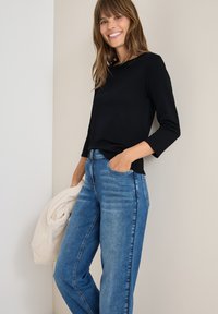 Schwarzes Langarmshirt kombiniert mit hellblauen geraden Jeans. Das Model hält eine cremefarbene Decke und fördert einen lässigen, entspannten Look.