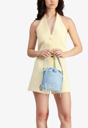 Borsa a tracolla intrecciata blu chiaro con chiusura a coulisse, tracolla removibile e dettaglio hardware circolare. Modella che indossa un vestito giallo pallido.