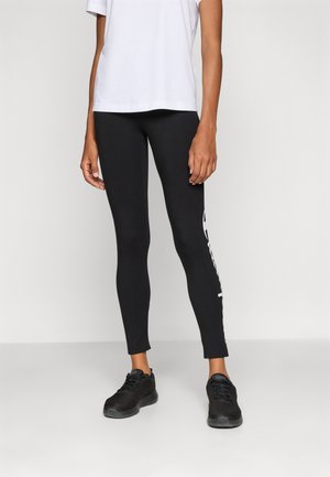 Svarte leggings med slim fit, som har hvit merkevare på siden. Kombinert med en løs hvit t-skjorte og svarte sneakers.