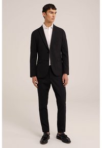 Un costume noir avec une veste cintrée et un pantalon slim, associé à une chemise blanche et des chaussures de ville noires, sur un fond clair.