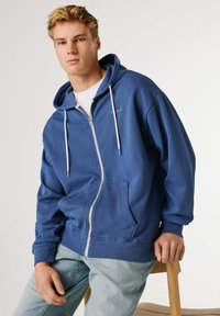 Niebieska bluza z zamkiem, wykonana z fakturowanego materiału, z kieszenią kangurką, kapturem ze sznurkiem, ściągaczami na rękawach i subtelnym logo.