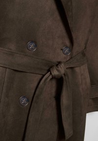 Close-up van een donkerbruine suède jas met een gestrikte riem en vier zichtbare zwarte knopen aan de voorkant.