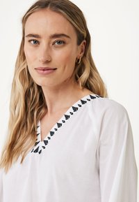Camicia bianca con scollatura nera ricamata, design a forma di V, tessuto morbido e maniche fluide. Cuciture minimaliste e dettagliate.