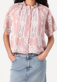 Vaaleanpunainen lyhythihainen paita, jossa on kukkakuvioinen pitsikoristelu, napit edessä ja aaltoileva helma. Yhdistetty siniseen farkkuhameeseen.