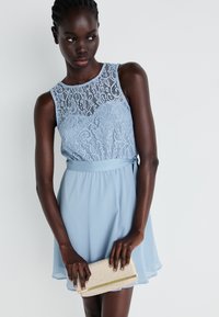 Svetlo modra obleka z čipkastim zgornjim delom, brez rokavov in razširjenim krilom. Manekenka drži bleščečo torbico z zlatimi poudarki.