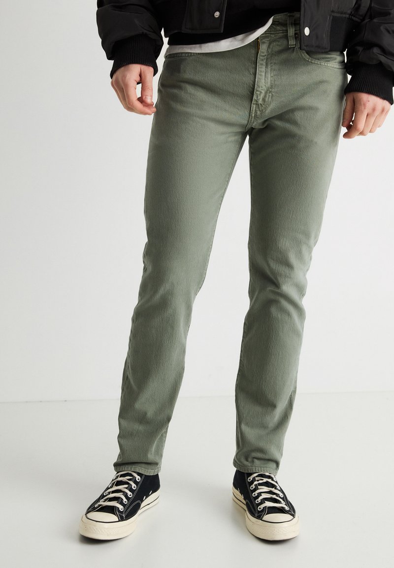 Άνδρας που φοράει ελαφρώς στενά χακί πράσινα τζιν, μαύρα και λευκά παπούτσια υψηλού τύπου και μαύρο μπουφάν, στέκεται σε λευκό πάτωμα.