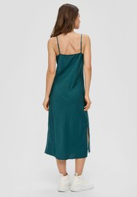 Sukienka bez rękawów w odcieniu zielono-niebieskim, o gładkiej fakturze, z bocznymi rozcięciami i cienkimi, regulowanymi ramiączkami. Noszona z białymi trampkami typu high-top.