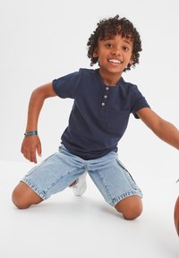 Camisa henley de manga corta azul marino con tres botones y un bolsillo, combinada con pantalones cortos de cargo de mezclilla clara y zapatillas blancas.