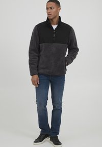 Fleece pulli i mørkegrå med en sort panel, kvart-zip front, sidelommer og ribbede manchetter. Båret med blå jeans og sorte sneakers.