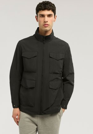 Mężczyzna w czarnej kurtce zapinanej na zamek z czterema kieszeniami na patki, stojący na tle jednolitego, jasnoszarego tła.