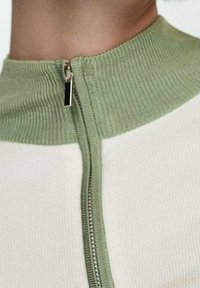 Haut beige clair avec un col côtelé vert haut et une fermeture éclair centrale. Le tissu semble lisse et extensible, avec des accents de couleur contrastants.