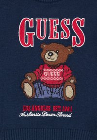 Brun dukkebjørn iført en rød genser med "GUESS" og blå jeans på marineblått stoff, med teksten "GUESS", "Los Angeles EST. 1981" og "Authentic Denim Brand".