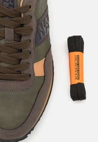 Baskets vert olive avec des accents en suède et cuir, lacets marron, et une étiquette de logo. Lacets de remplacement noirs avec une étiquette orange sur le côté.