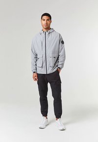 Giacca con cappuccio grigio chiaro con due tasche frontali, dettagli neri e chiusura con zip, abbinata a pantaloni cargo neri e scarpe da ginnastica bianche.