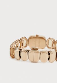 Montre dorée avec un bracelet en maillons géométriques, présentant des formes octogonales et rectangulaires. Le dos est gravé et possède une couronne ronde.