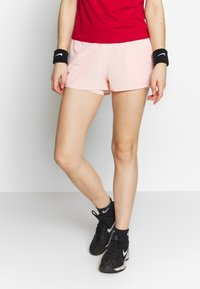 Pantalones cortos deportivos de color rosa claro con una cintura elástica, logo negro de Nike en el lateral. Combinados con calzado negro y muñequeras. Camiseta roja encima.